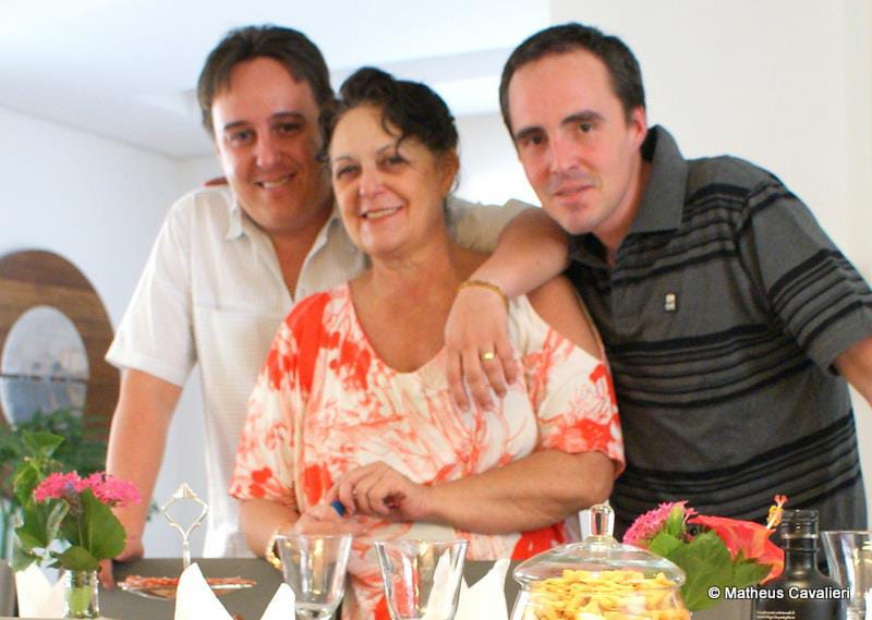 Família reunida à mesa — mãe entre os dois filhos, flores e comida