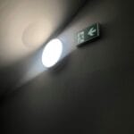 Illuminated emergency exit sign with light casting shadows on a dark wall.